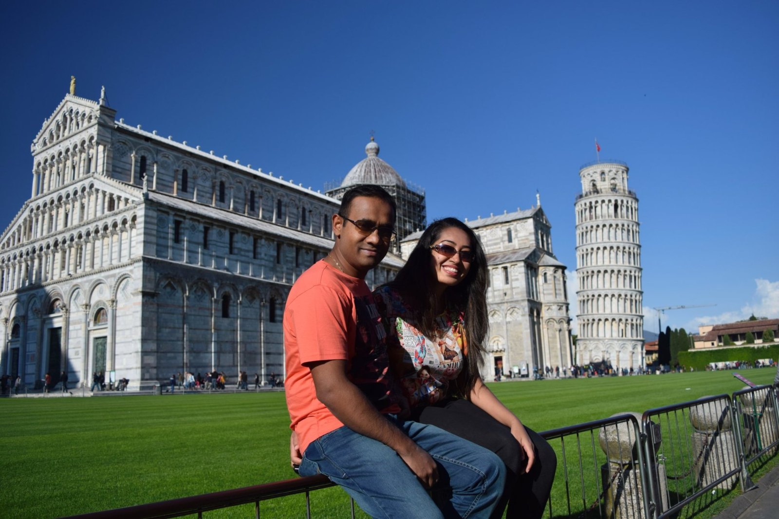 Meenu and Praddy sitting on a railing on the lawn, with Pisa's cathedral and the Leaning Tower behind them