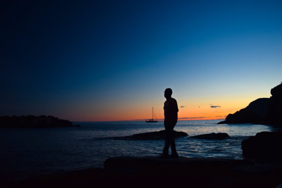 Praddy's silhouette on the rocks against the last orange light, with a sailing boat on the sea