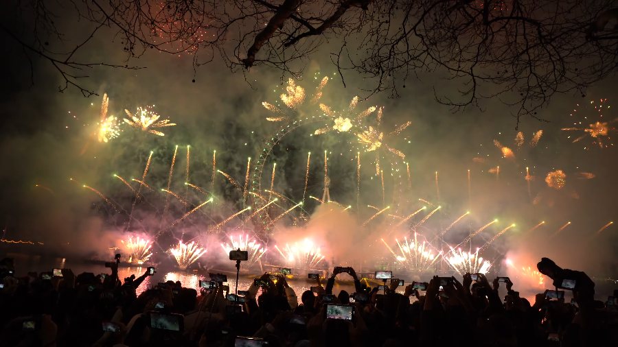 Gold shells bursting over a row of crossing gold fountains along the river, with the crowd filming