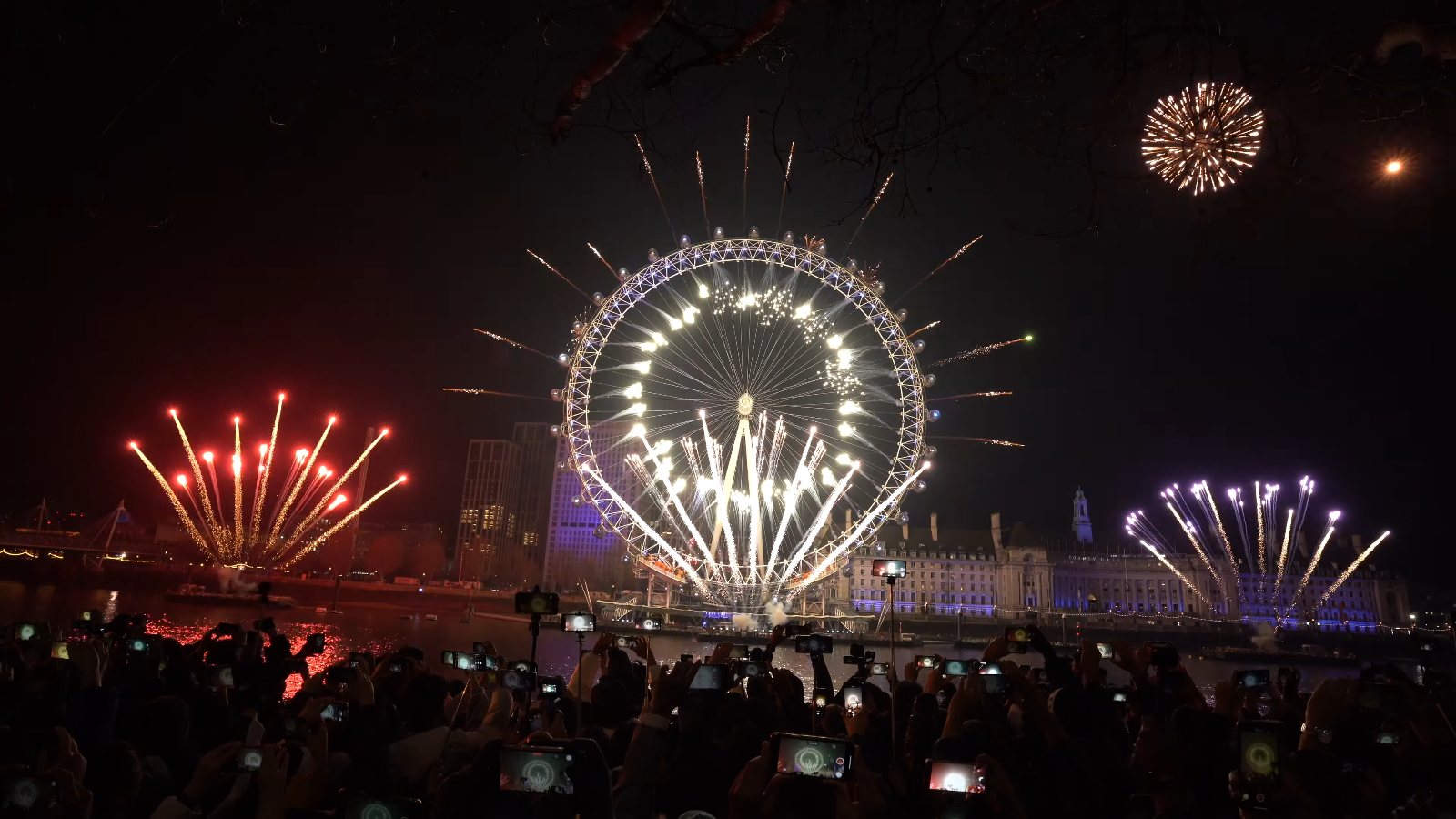 The London Eye lit up white at midnight, with sparks shooting out from its rim and red and lilac fans on either side