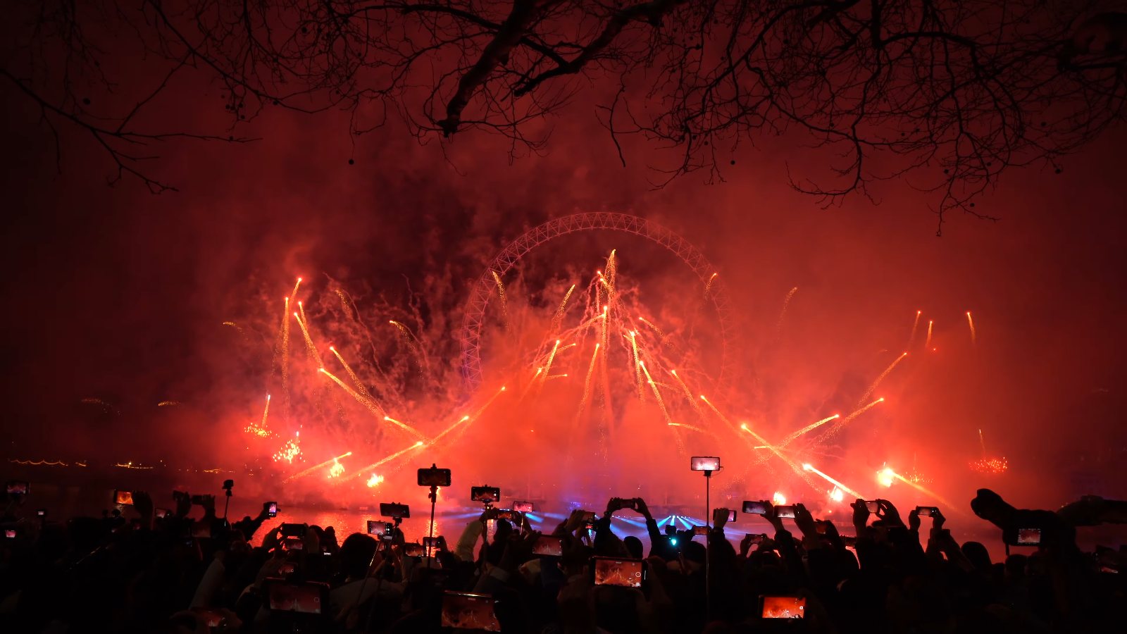 Red fans of sparks criss-crossing in red smoke over the river, with phones held up in the crowd