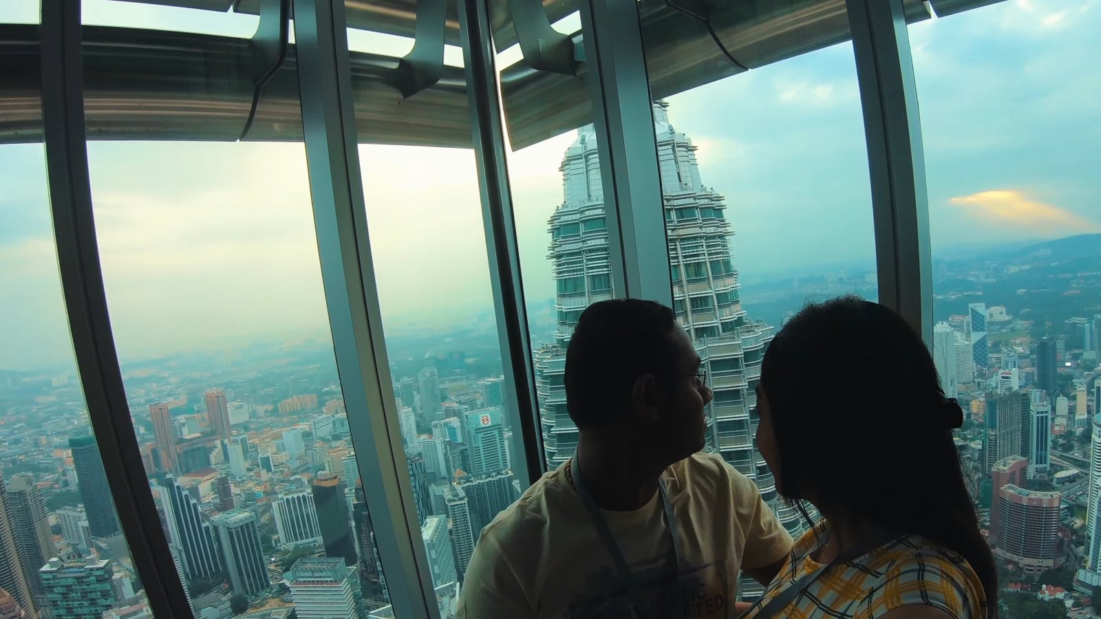 Praddy and Meenu at the window of the Petronas Towers' 86th-floor observation deck, with the other tower right outside the glass