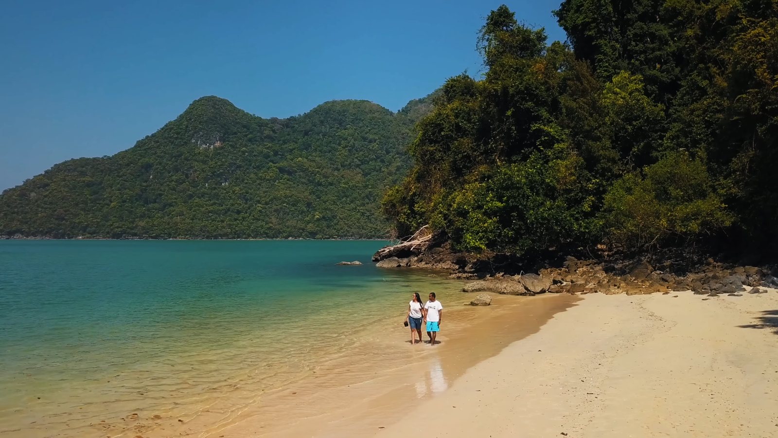 Drone view of the two of us at the water's edge on a white-sand beach, with a jungle headland and a mountainous island behind