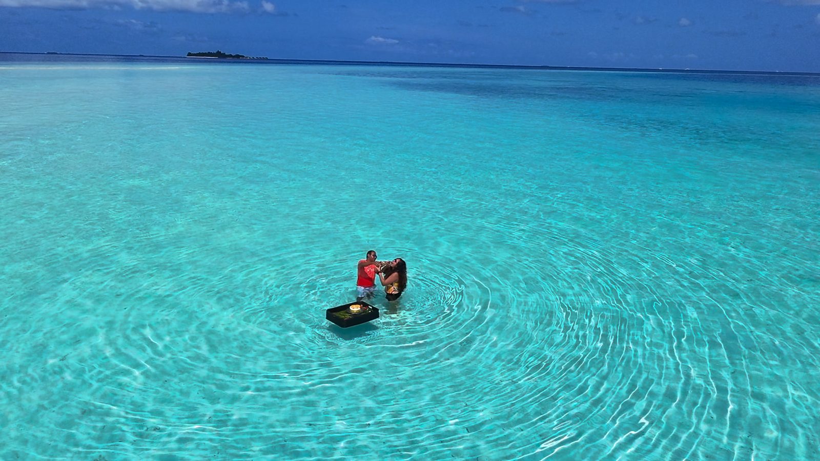 Meenu and Praddy toasting waist-deep in the lagoon beside a floating tray with a birthday cake