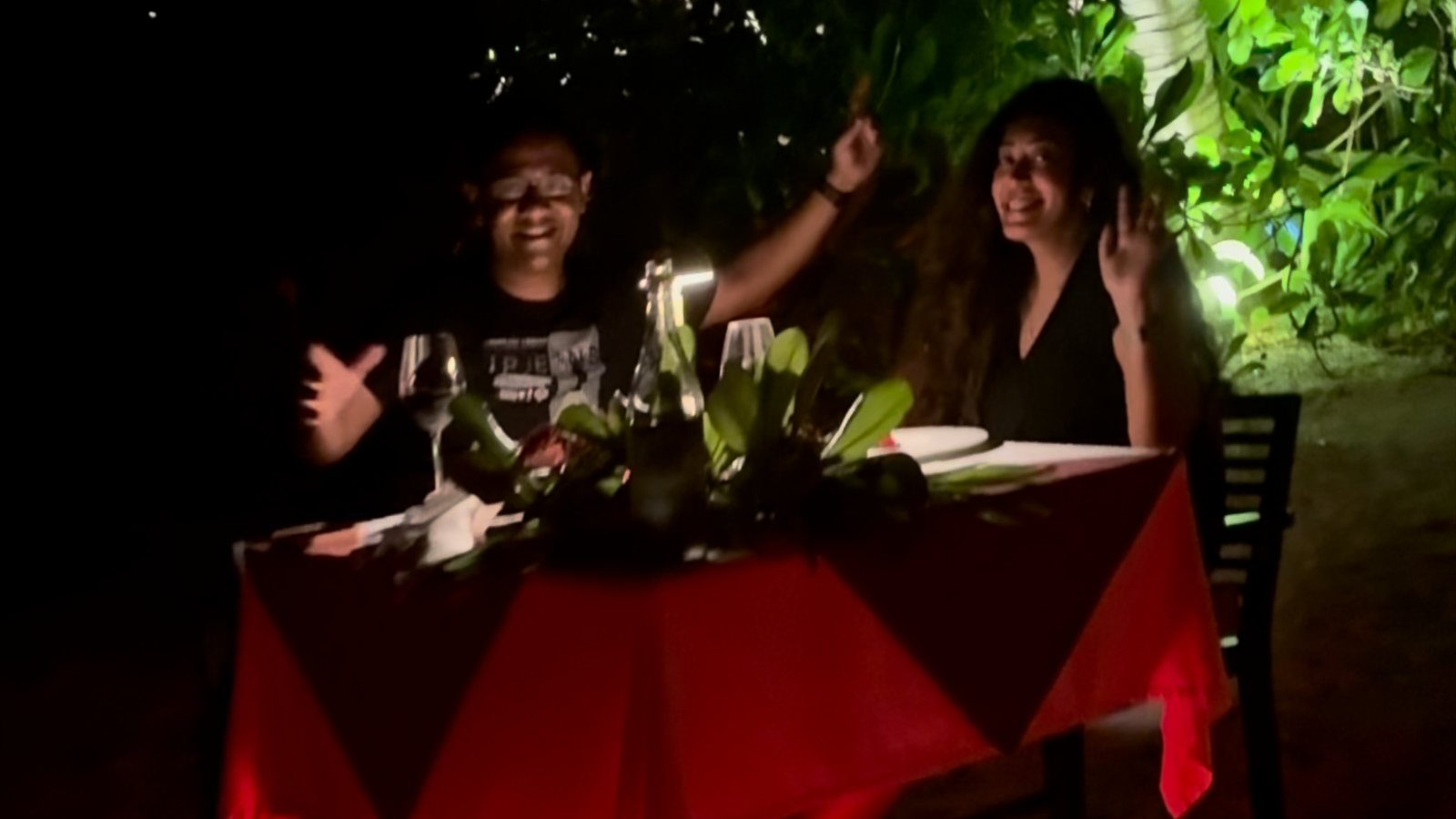 Meenu and Praddy waving from a candlelit table with a red cloth in the garden at night