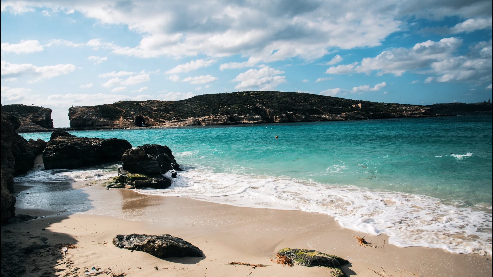 A little beach of pale sand and dark rocks, with turquoise water and a low island across