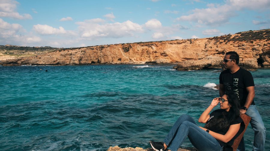 Praddy standing and Meenu sitting on the rocks above the turquoise lagoon, with bare orange cliffs across the water