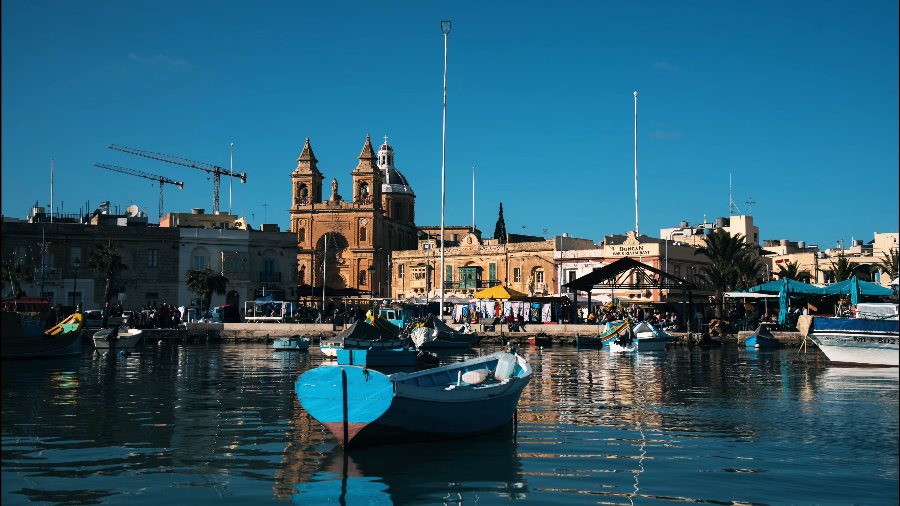 Painted luzzu fishing boats in Marsaxlokk harbour, in front of the church's twin bell towers