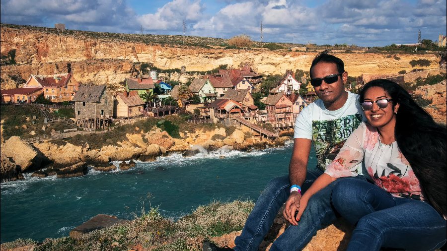 Meenu and Praddy on the rocks above Anchor Bay, with the painted wooden houses of Popeye Village behind