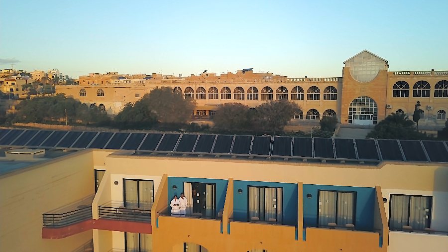 The two of us in white robes on our balcony, with solar panels on the roof and the arched Corinthia St George's behind