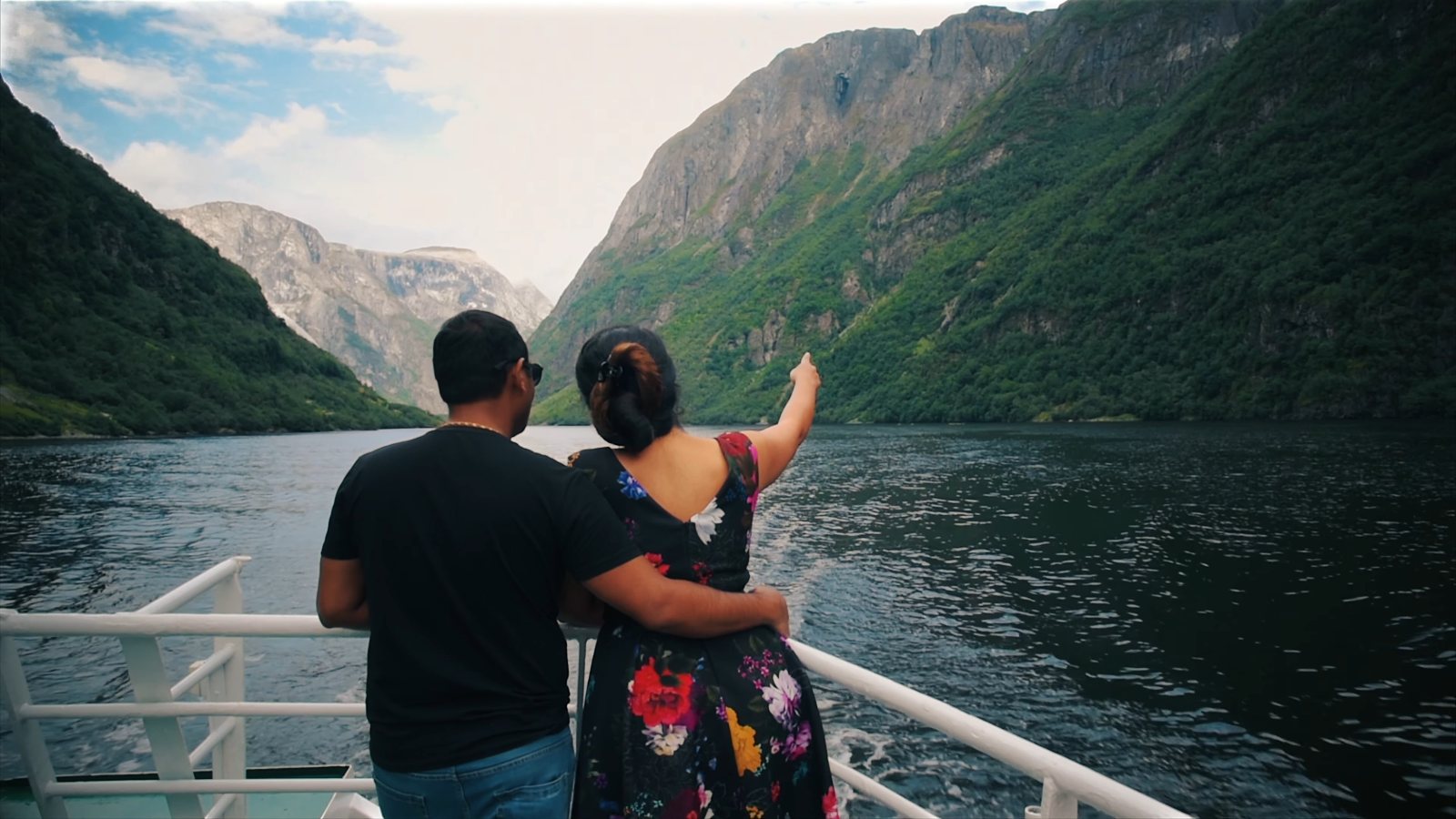 Praddy and Meenu at the back rail of a boat, Meenu pointing up at the fjord's steep green walls