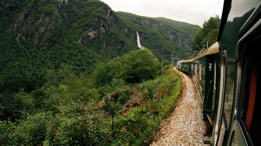 The green Flåm Railway train curving along a wooded mountainside, with a waterfall behind