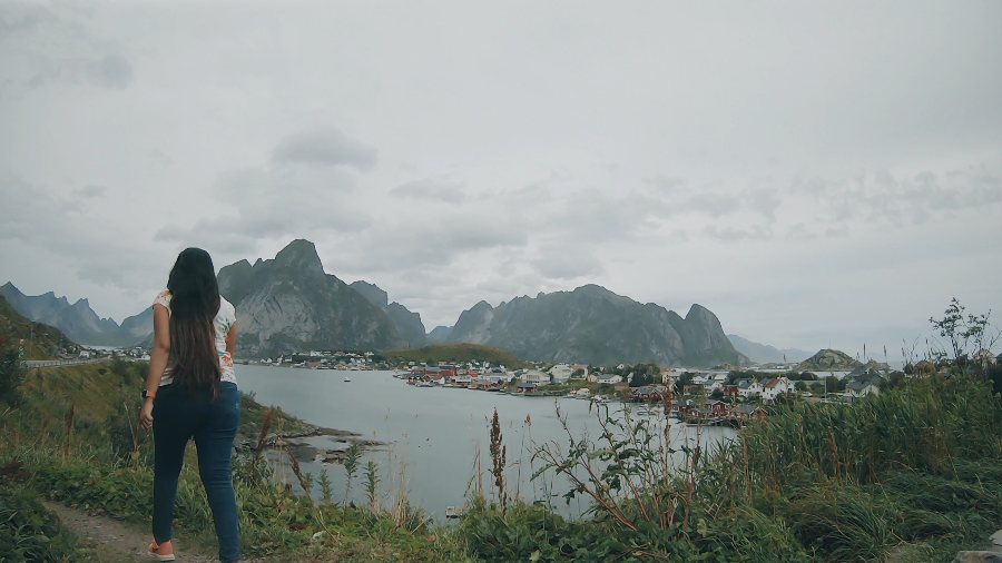 Meenu walking toward a village on the water under jagged peaks