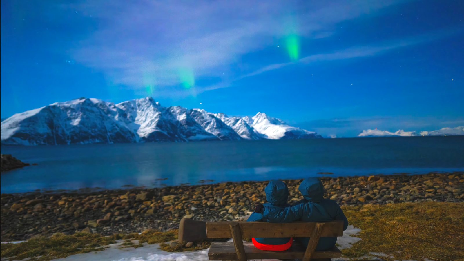 The two of us on a bench on the shore, with the aurora over the snowy peaks across the water