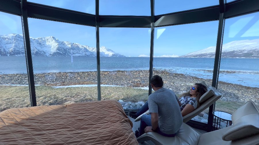 Praddy and Meenu on loungers in the glass-walled cabin, facing the fjord and mountains