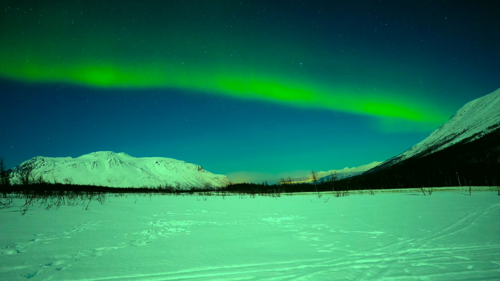 A green arc of aurora over a snowy plain and mountains