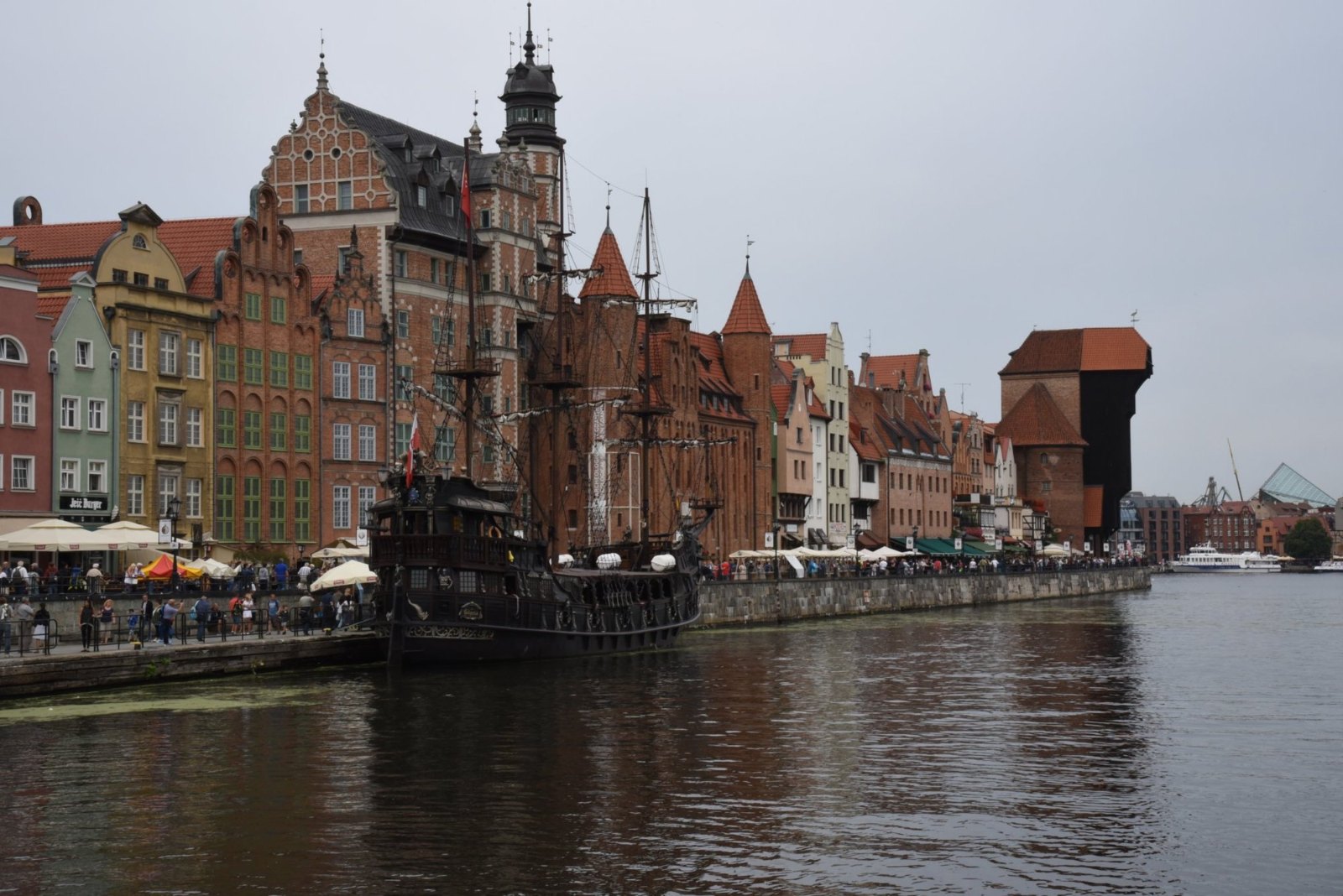 Gdańsk's waterfront on the Motława, with tall gabled houses, a black wooden galleon moored in front and the old Crane at the far end