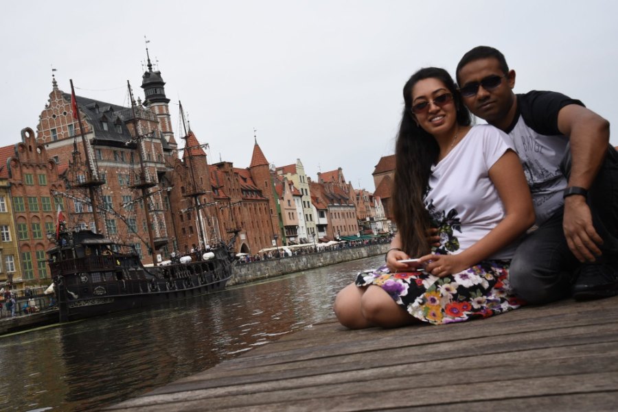 Meenu and Praddy sitting on a wooden boardwalk across the river from the galleon and the brick houses of the old town