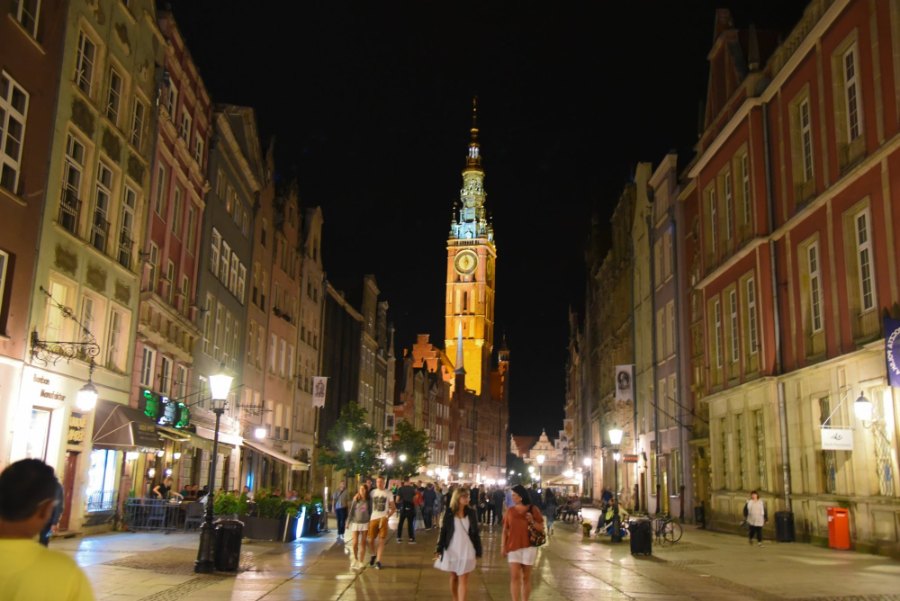 The old town's main street at night, busy with people, with the Town Hall tower lit up at the far end