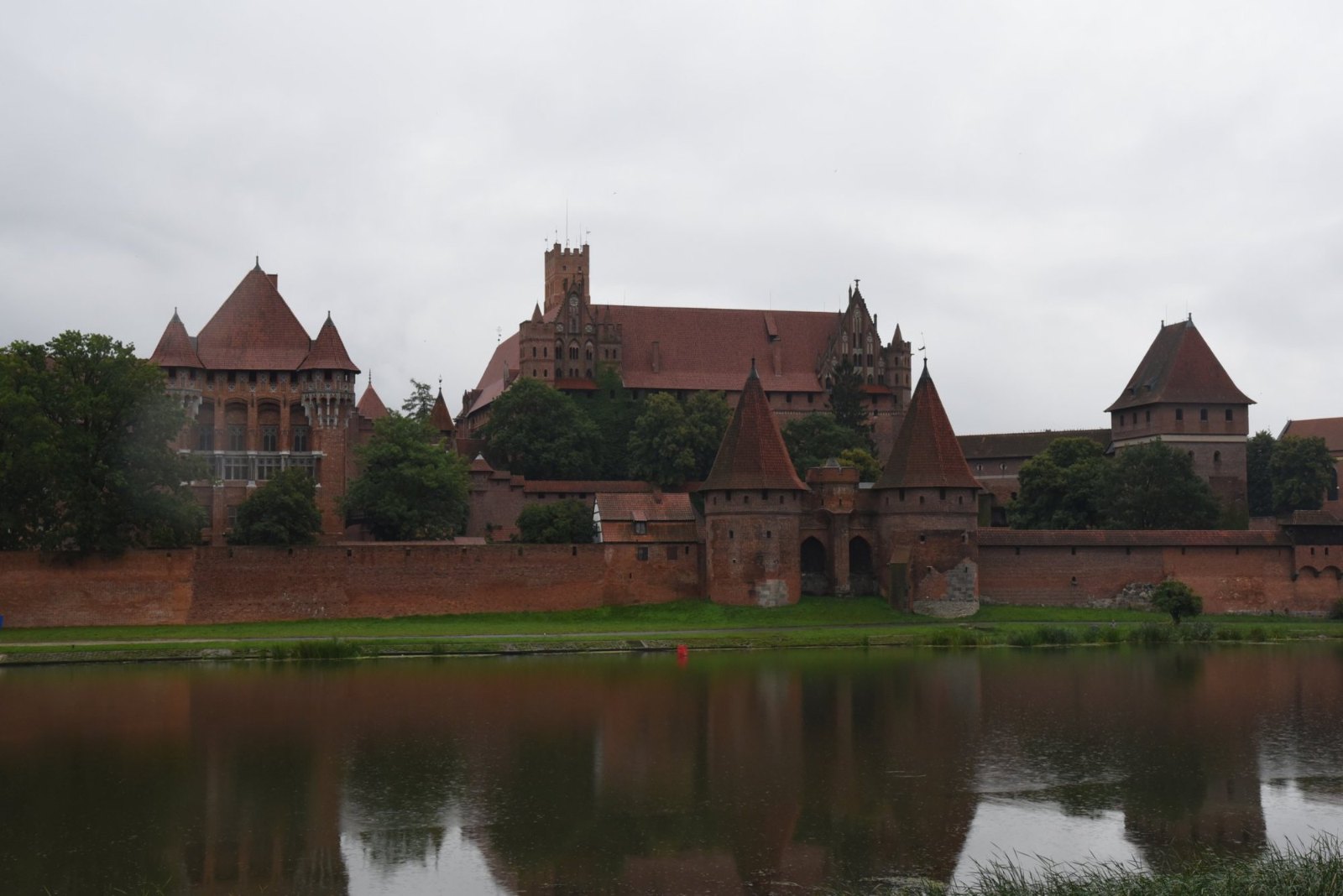 Malbork Castle's red-brick towers, roofs and walls across the river Nogat under a heavy grey sky, reflected in the water