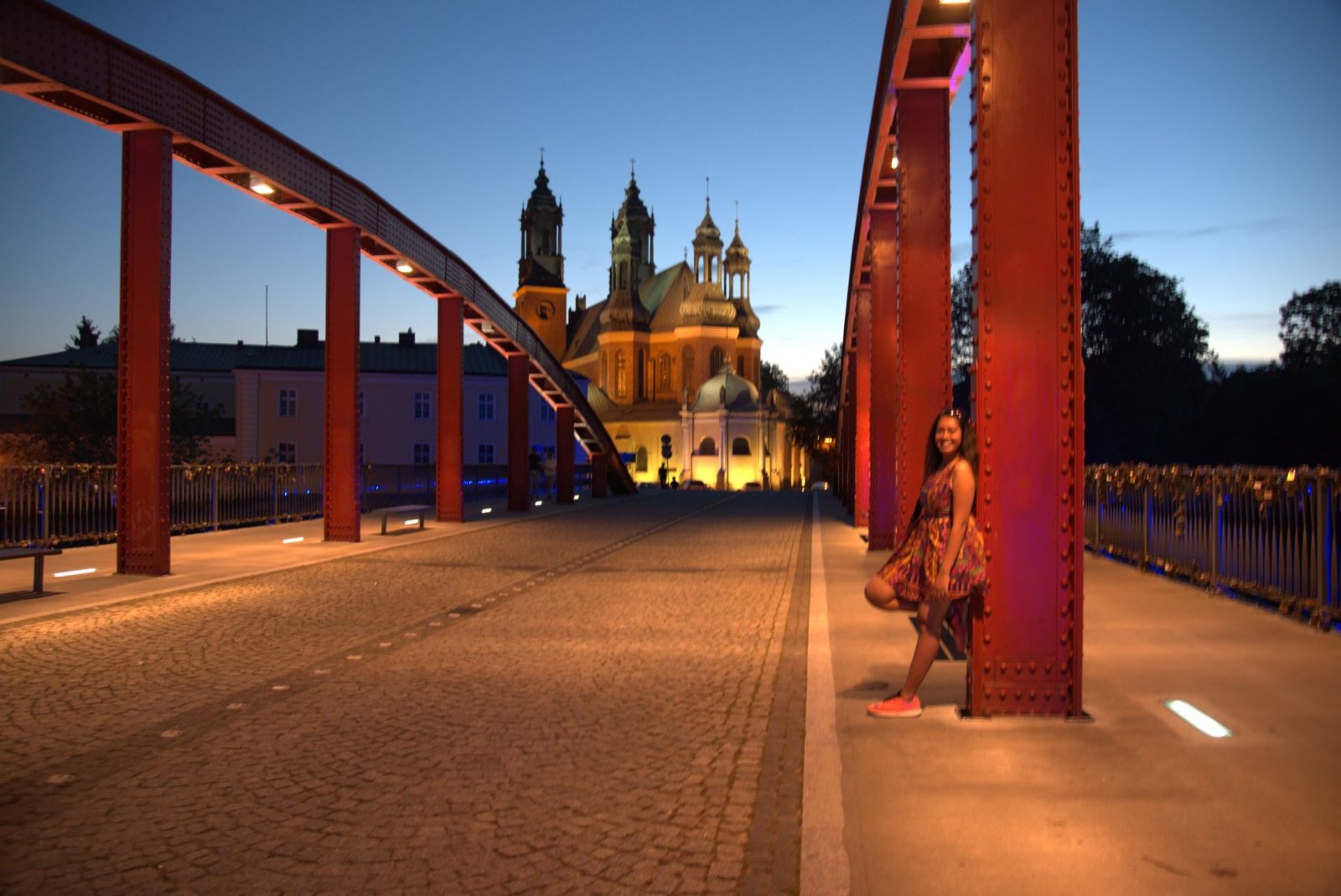 Meenu on the red-arched Jordan Bridge at dusk, with Poznań Cathedral floodlit gold beyond