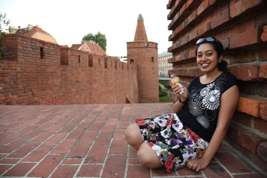 Meenu smiling with an ice cream, sitting against the brick walls of the Old Town by the Barbican's tower