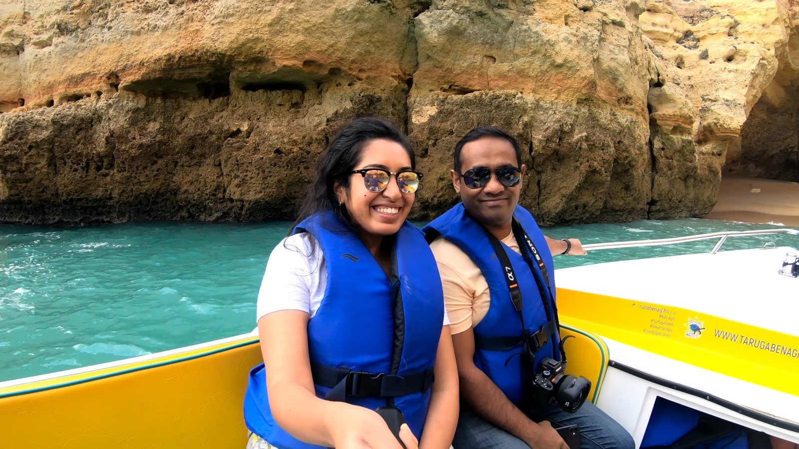 Meenu and Praddy smiling in blue life jackets on the yellow Taruga boat, with the cliffs behind