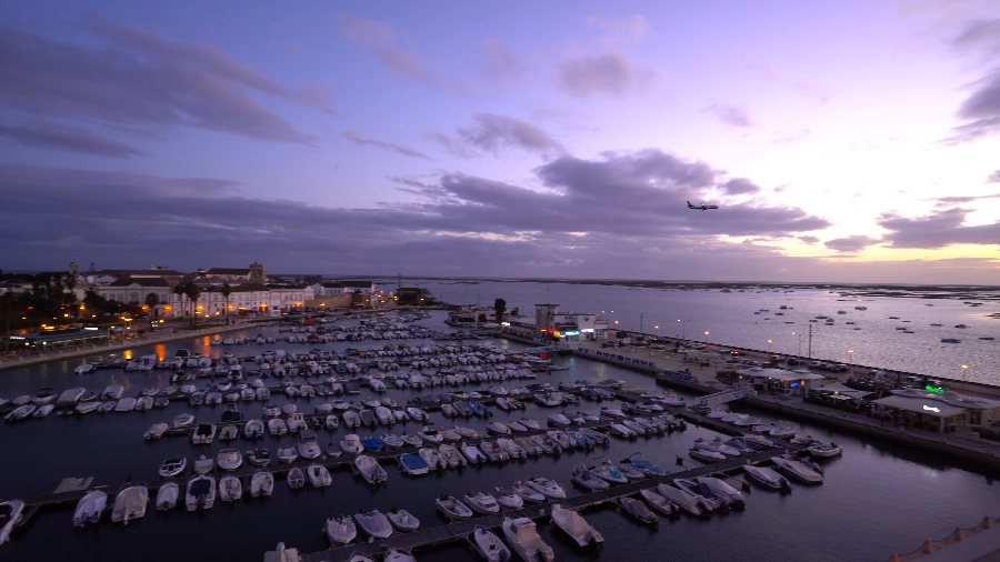 Faro's marina at dusk under a purple sky, with a plane coming in over the lagoon