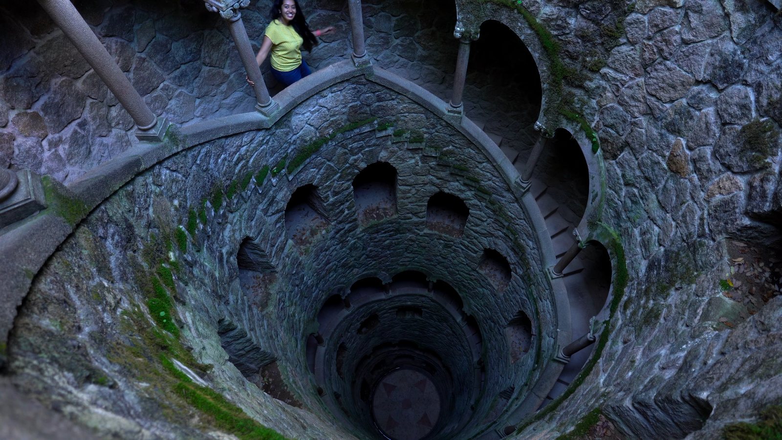Meenu waving from the top of the Initiation Well at Quinta da Regaleira, its spiral stairs dropping away below