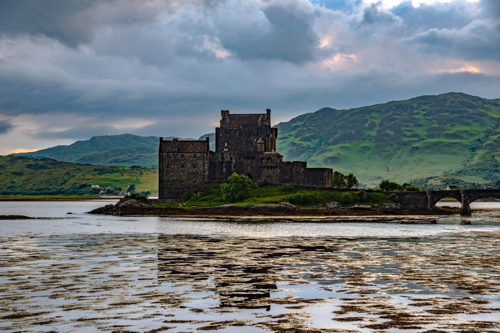 Eilean Donan Castle on its island, with the stone bridge and green hills behind and seaweed on the shore at low tide
