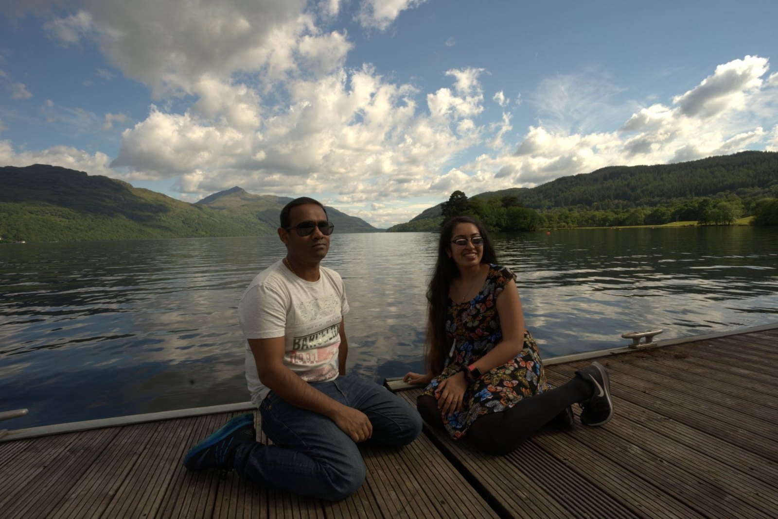 Praddy and Meenu sitting on a wooden jetty at Inveruglas, with Loch Lomond, its hills and big white clouds behind