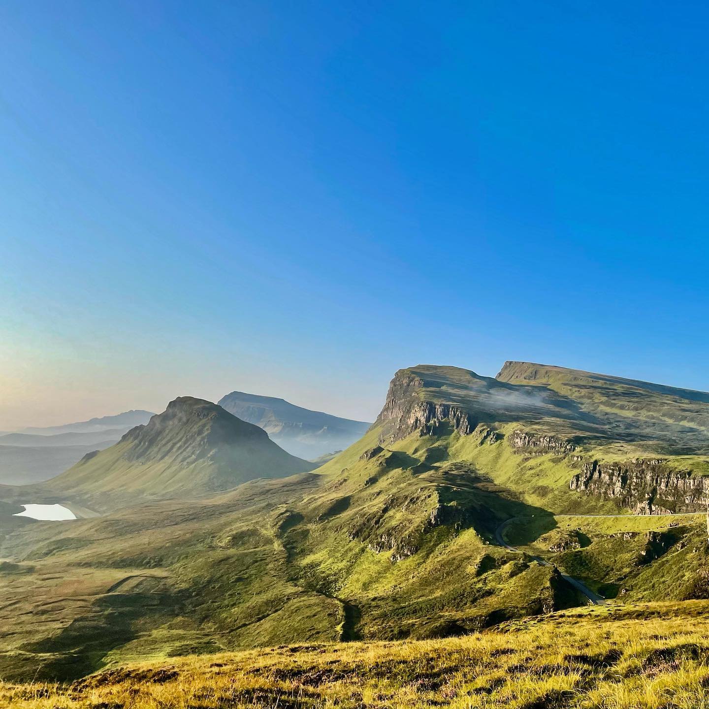 The Quiraing in early sun, its cliffs and green slopes falling away to a small loch and distant hills