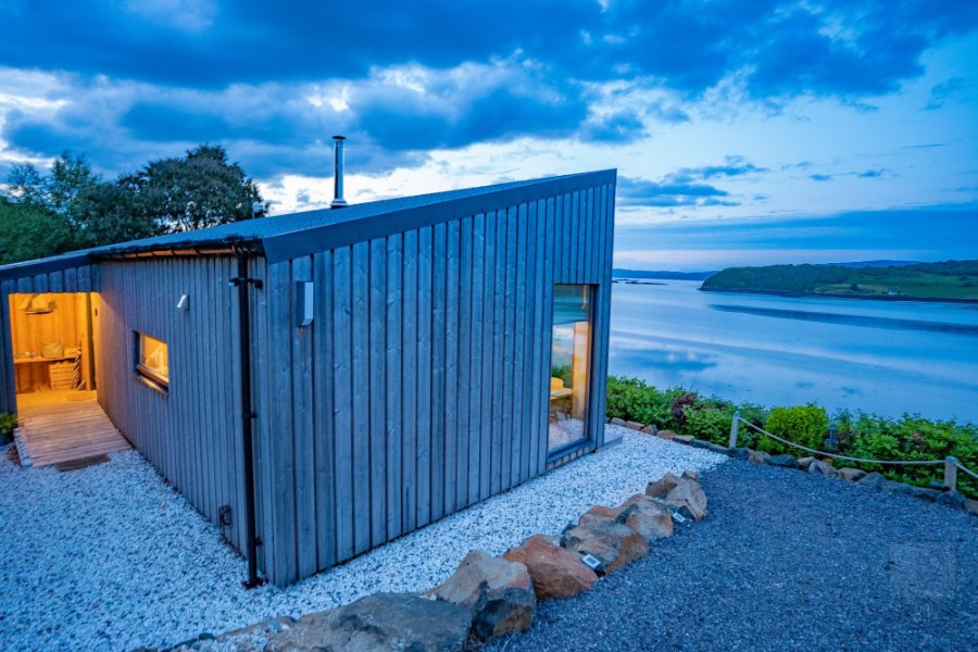 Air an Oir, a small cabin clad in grey timber, glowing at dusk above the water