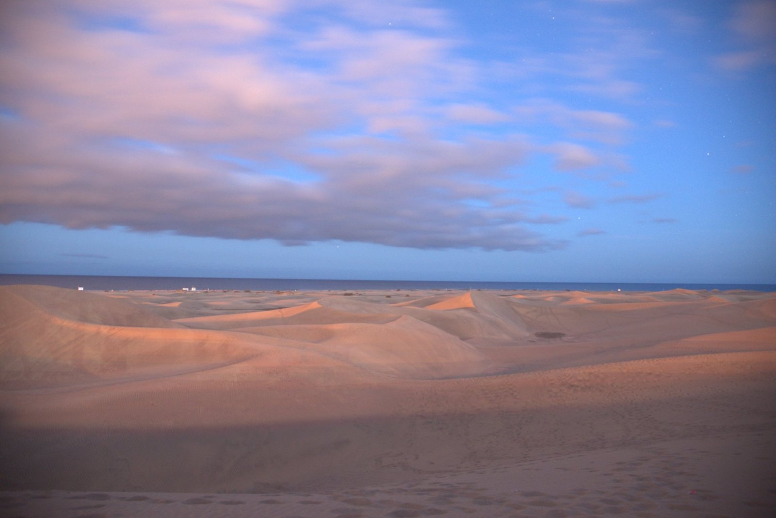 The Maspalomas dunes at dusk, ridges of sand under pink-tinged clouds, with the sea beyond
