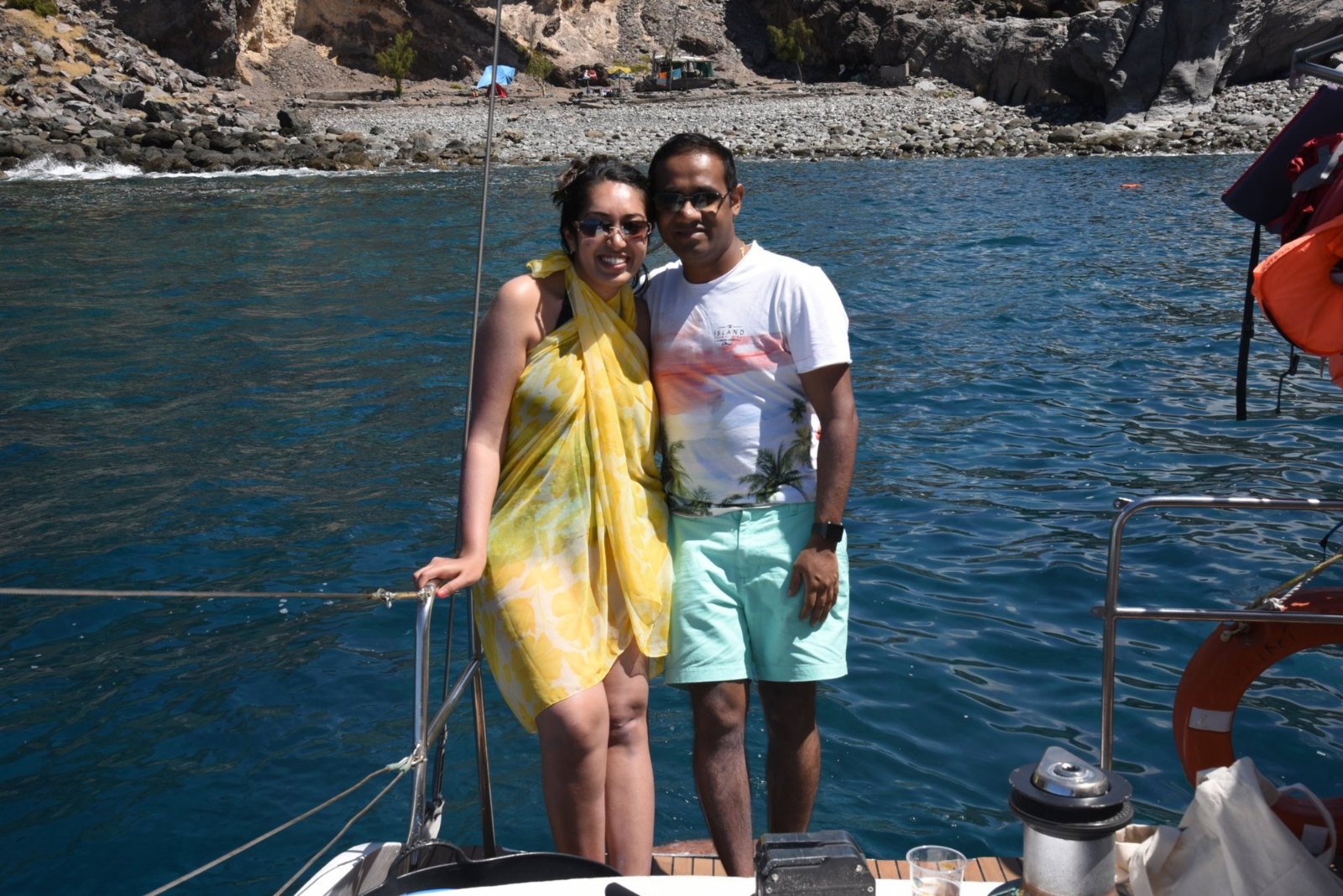 Meenu and Praddy on the catamaran's deck, with turquoise water and a pebble cove behind