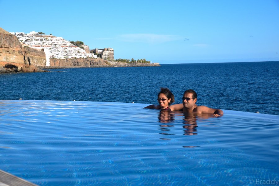 The two of us at the rim of an infinity pool that meets the ocean, below a cliff topped with white houses