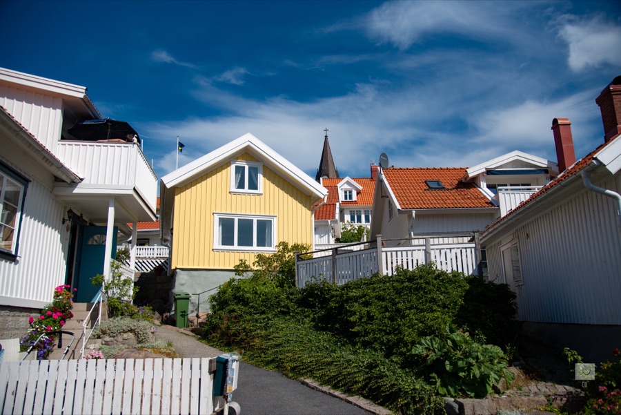 A yellow wooden house among white ones on a Fjällbacka lane, with the church spire behind