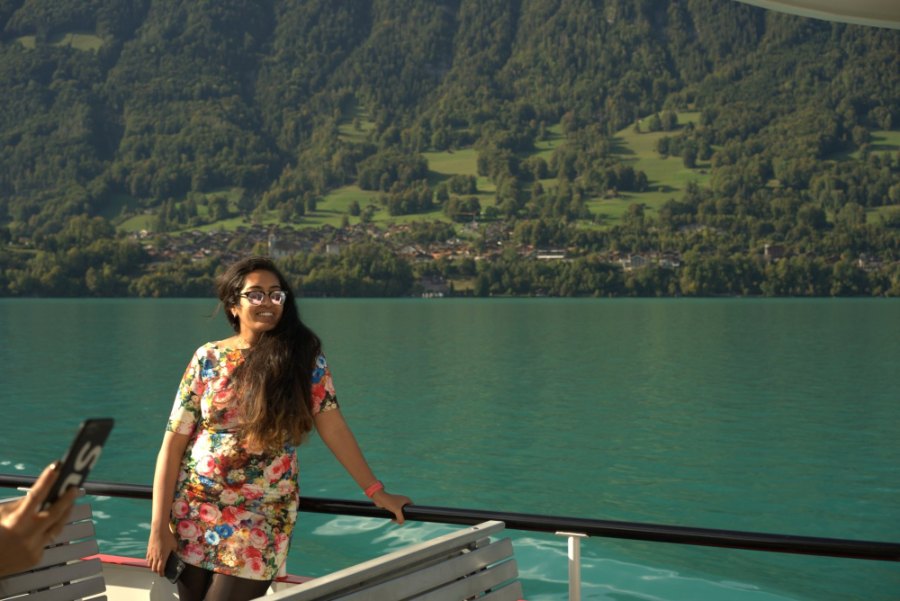 Meenu smiling at the boat's rail, with the turquoise lake and a wooded shore behind