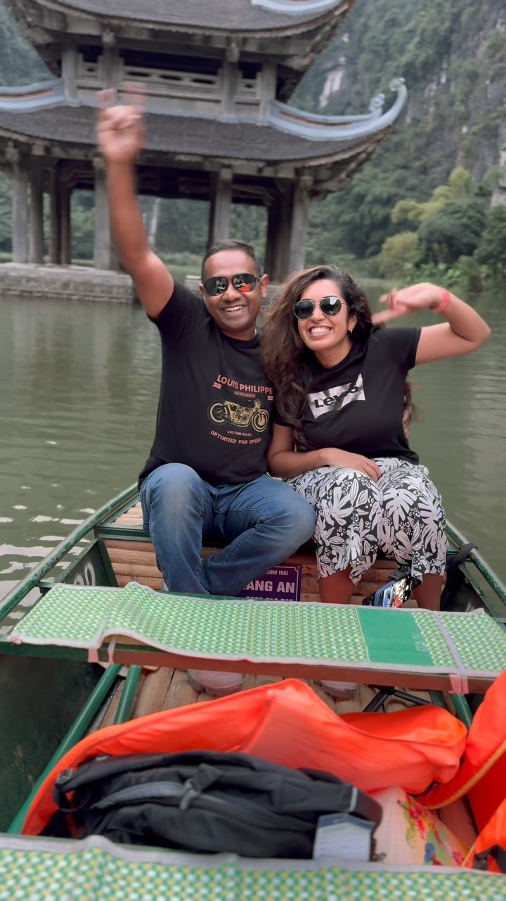Praddy and Meenu waving from a rowing boat at Tràng An, with a temple gate on the water behind