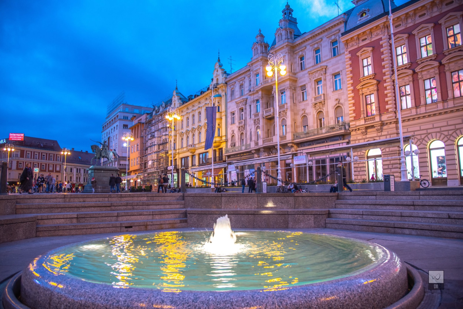 The fountain on Ban Jelačić Square at dusk, lit up in front of the glowing façades