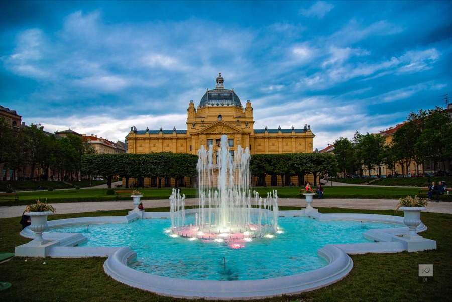 The yellow Art Pavilion under its dome, beyond a fountain lit pink and white at dusk